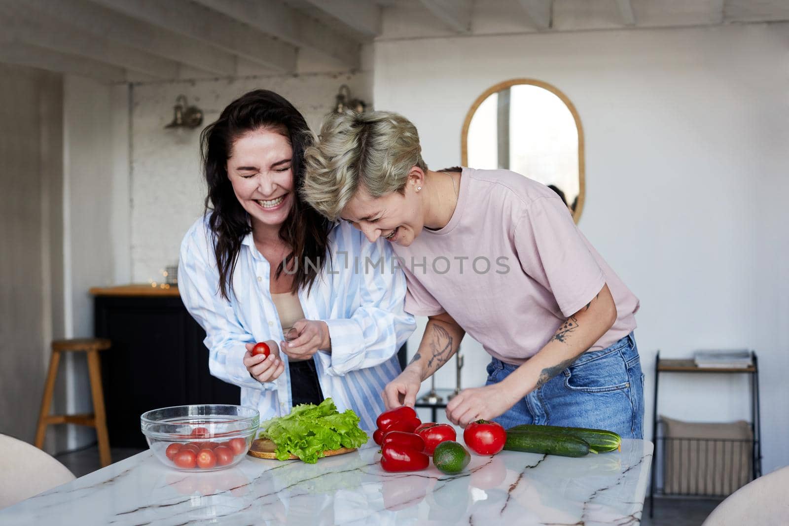 Cheerful young women in casual clothes laughing at joke while cooking healthy salad for lunch in kitchen at home