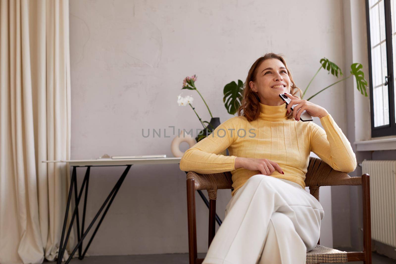 Young female in casual clothes holding mobile phone and dreaming happily while sitting on chair at home