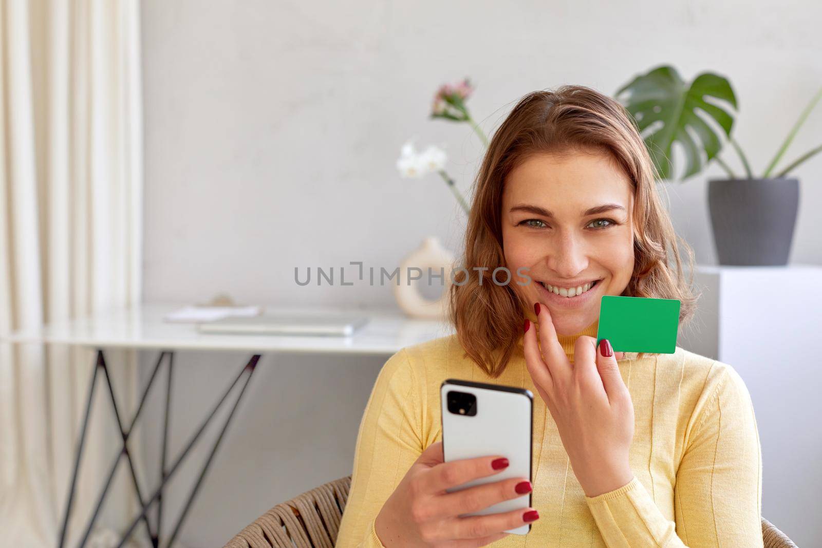 Delighted female with plastic card and smartphone making online purchase while looking at camera at home
