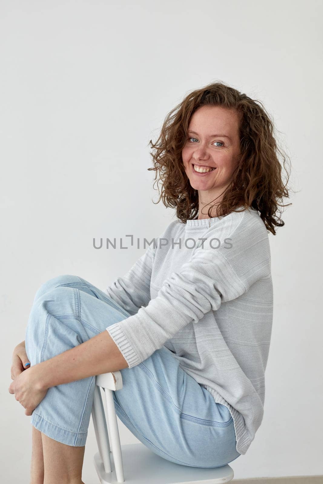 Side view of young female in trendy outfit sitting on wooden chair on white background and looking at camera