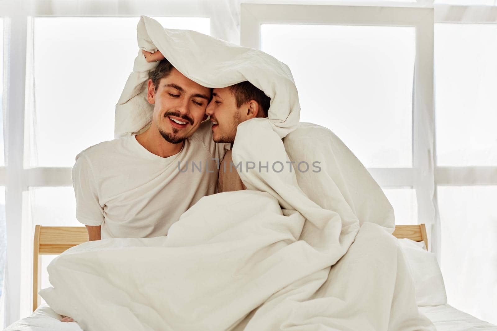 Happy young men wrapping in warm blanket and touching noses while resting on comfortable bed in morning in cozy bedroom