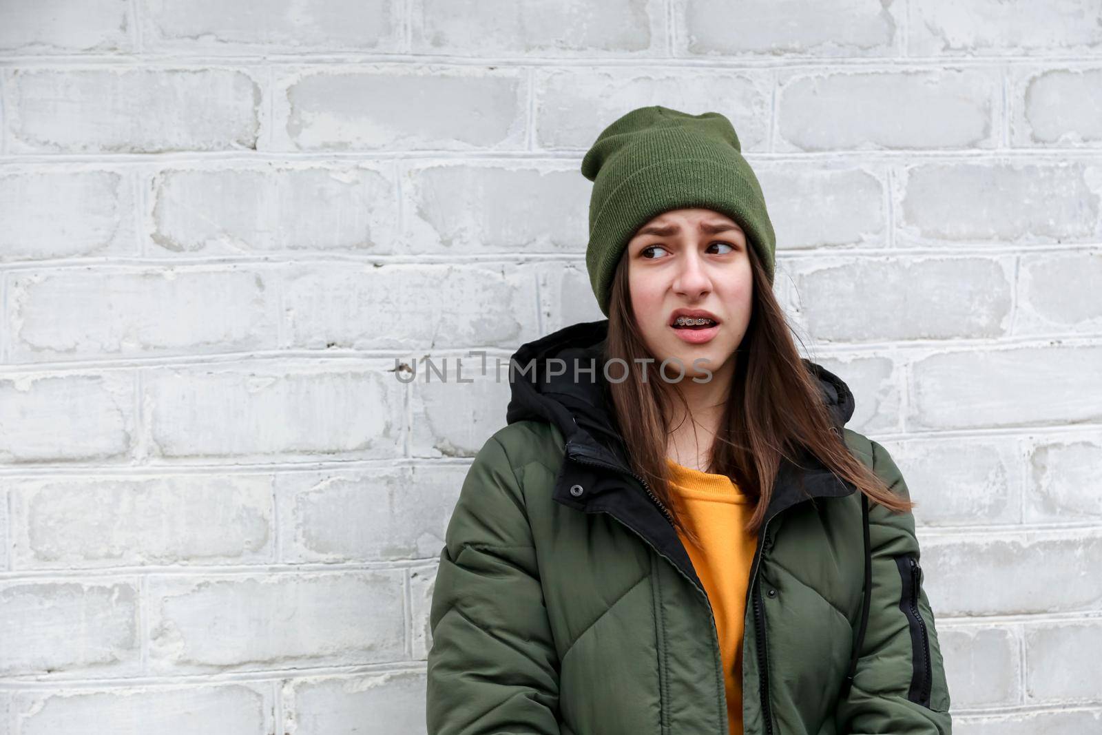 Portrait of a beautiful incredulous girl with braces in a yellow sweater and khaki hat that stands near a white brick wall. The concept of emotions. by lunarts