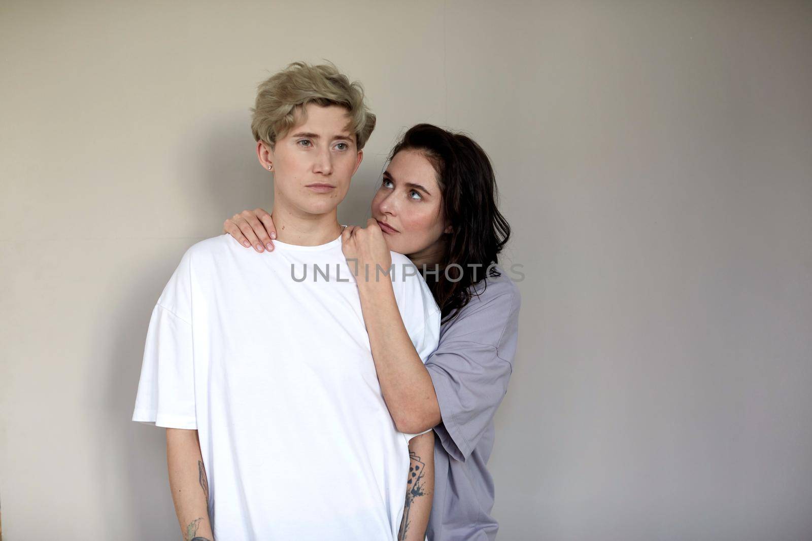 Young brunette embracing and looking at androgynous woman with short hair against gray wall