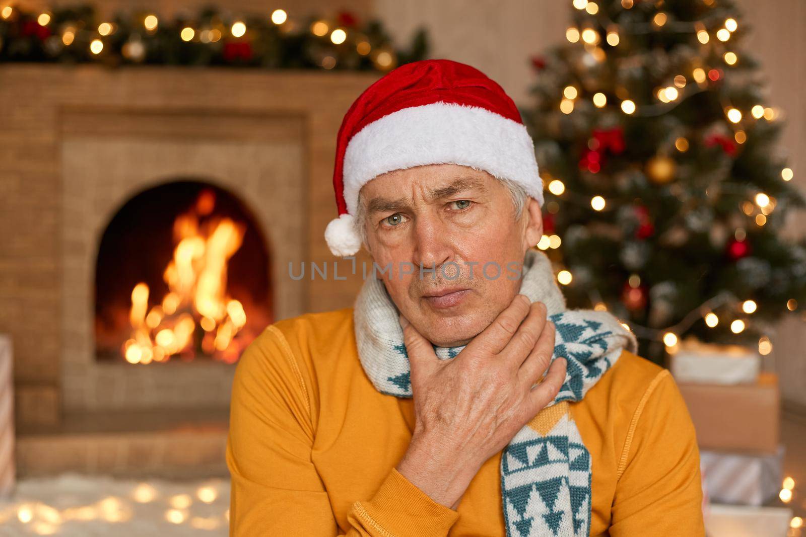 Sick man in orange sweater, scarf and Christmas hat suffering from sore throat, keeping hand on neck, posing on background of fireplace and fir tree, spends new year eve ant home.