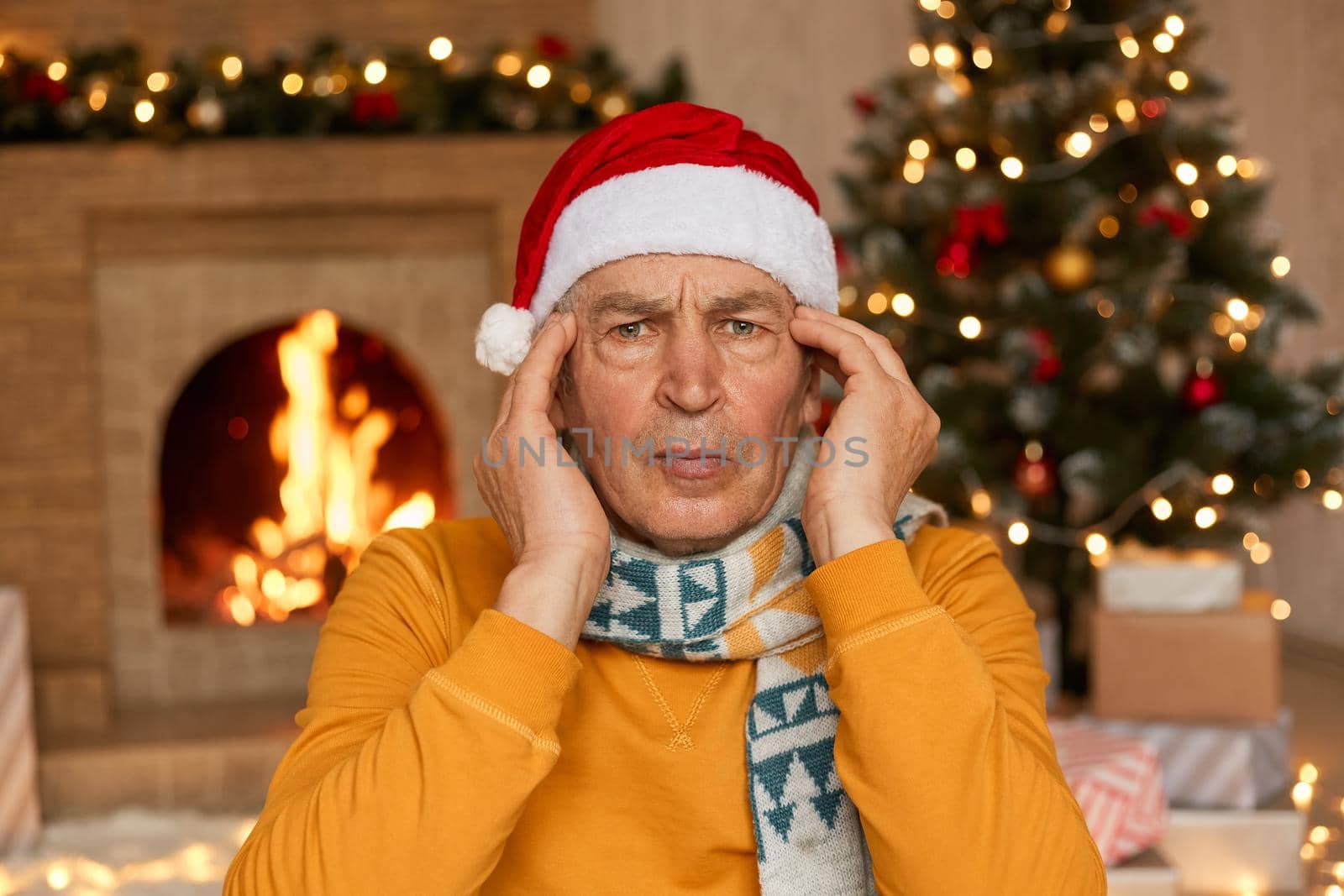 Senior male in Santa hat looking at camera and keeping hands on temples, suffering from headache, posing indoor near fireplace and xmas tree, being ill during new year eve.