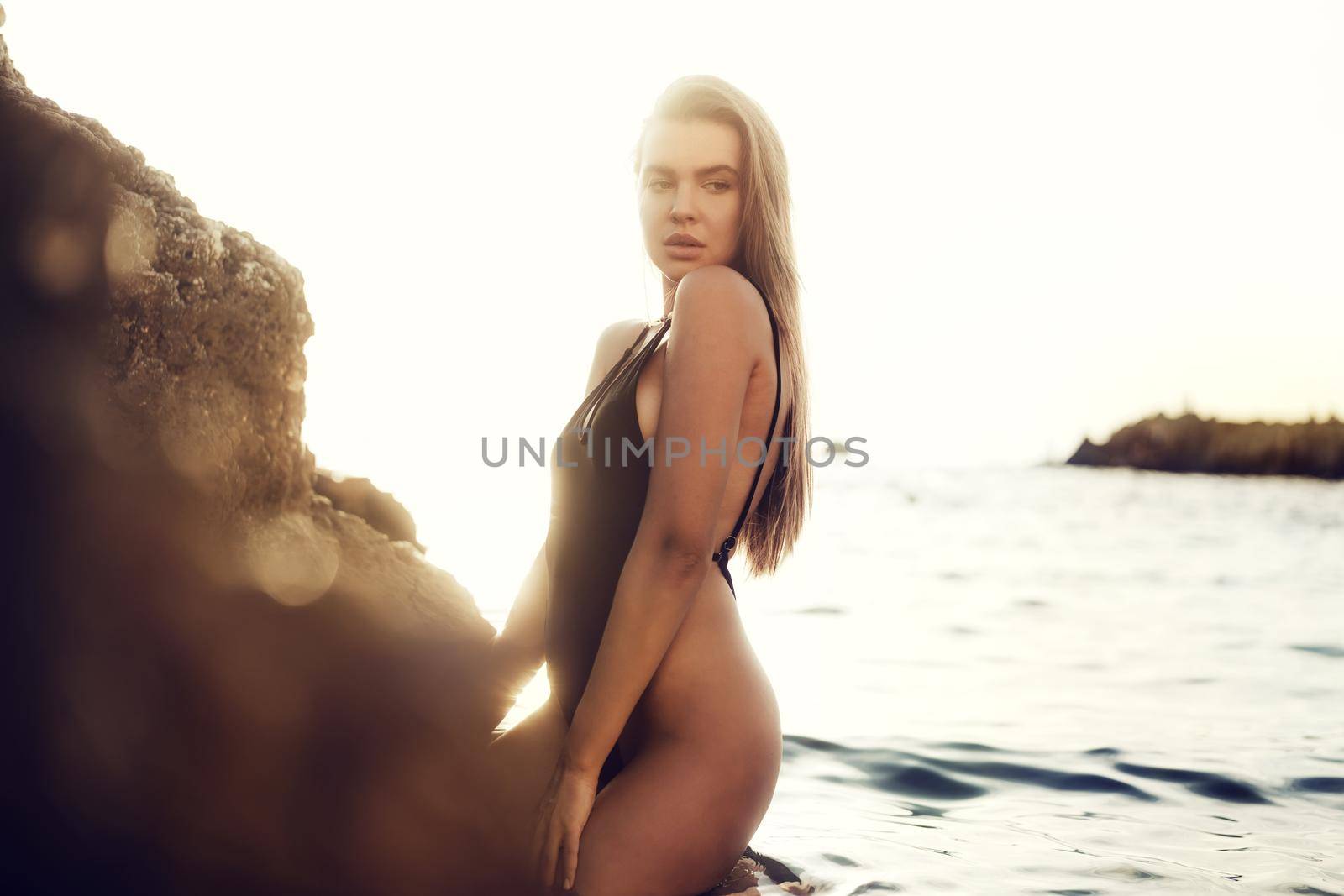 Young beautiful sexy female model in black bikini standing in sea water near huge rock