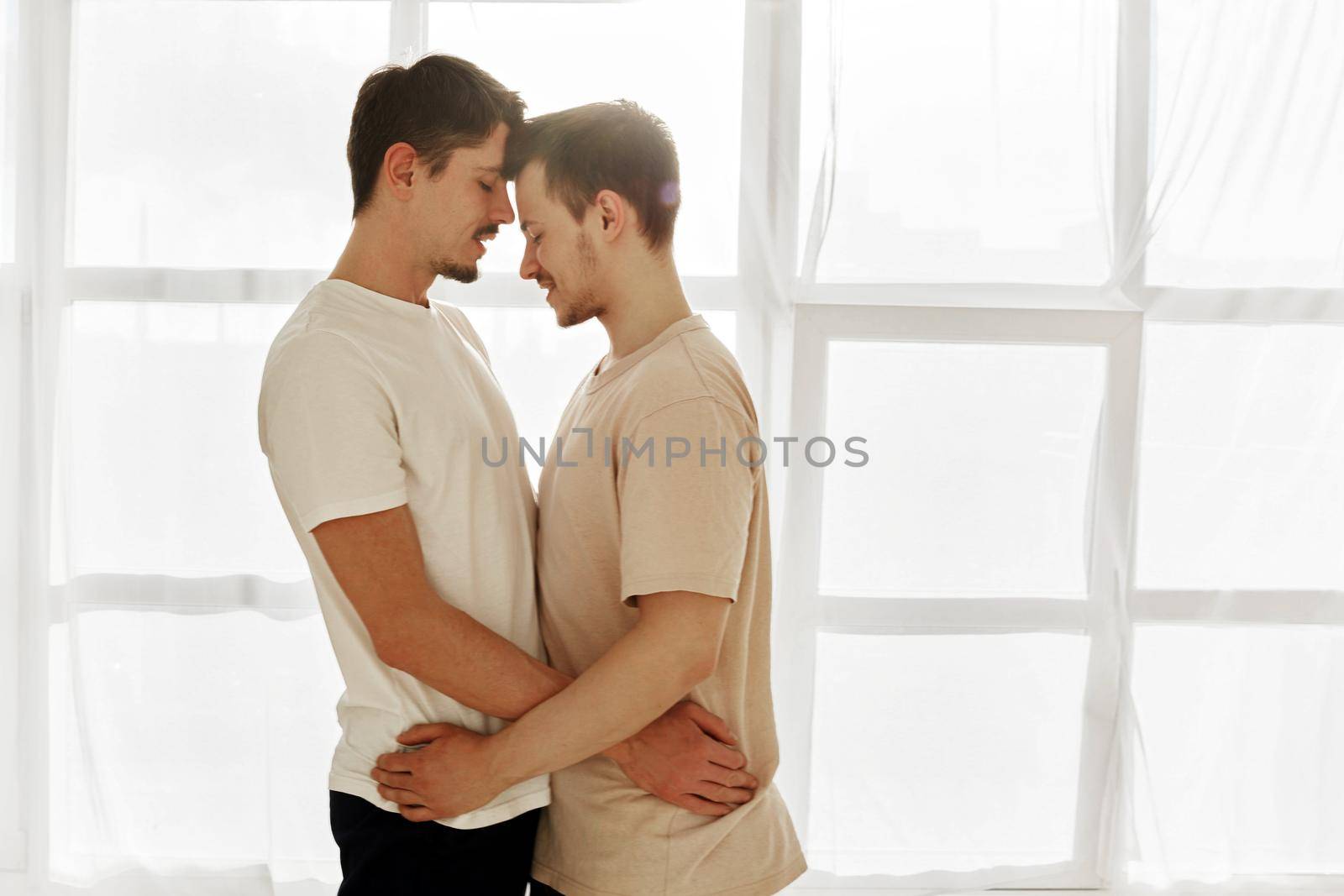 Side view of loving gay men in white t shirt leaning with foreheads and embracing against window
