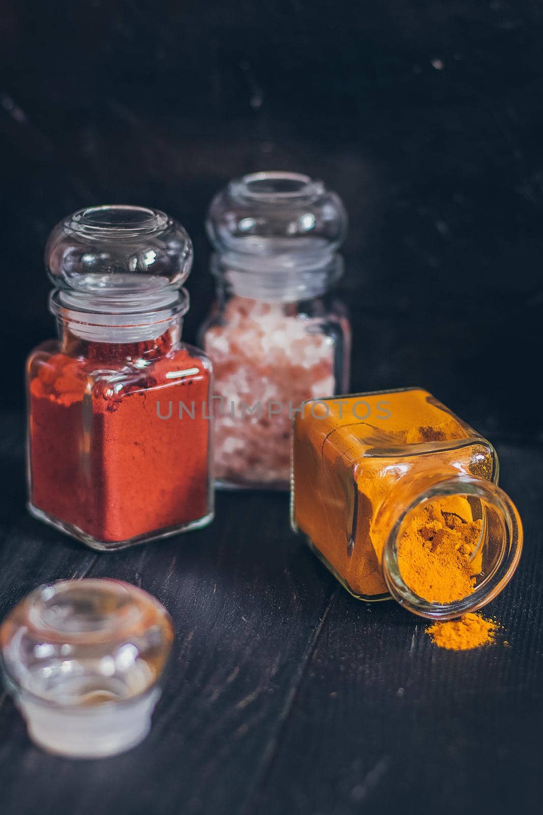 A set of spices in small jars with cork lids. Pink salt, ginger, turmeric, masala, paprika, smoked paprika. Spices on a black surface