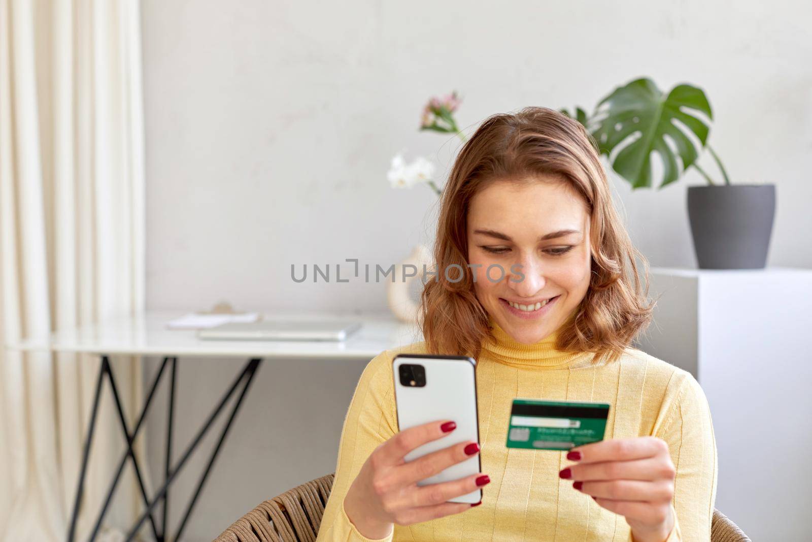 Delighted female with plastic card and smartphone making online purchase at home. Smiling woman paying by card for online purchase by phone