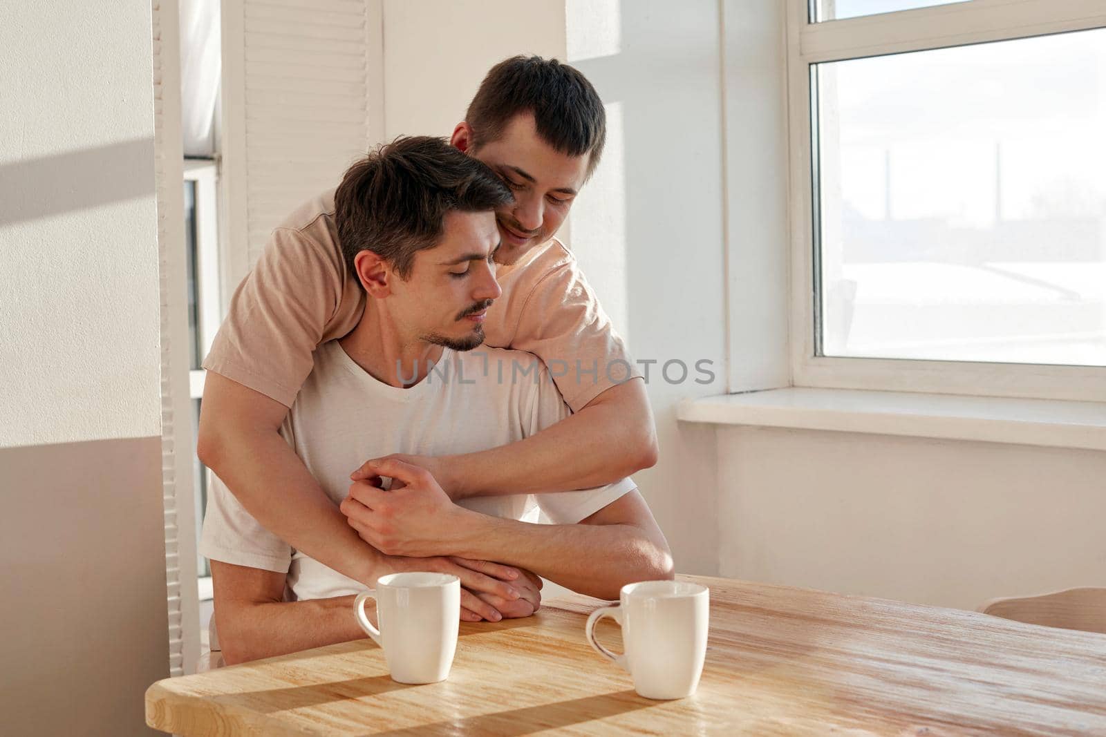 Happy young man embracing boyfriend from behind while resting at table during breakfast in morning at home