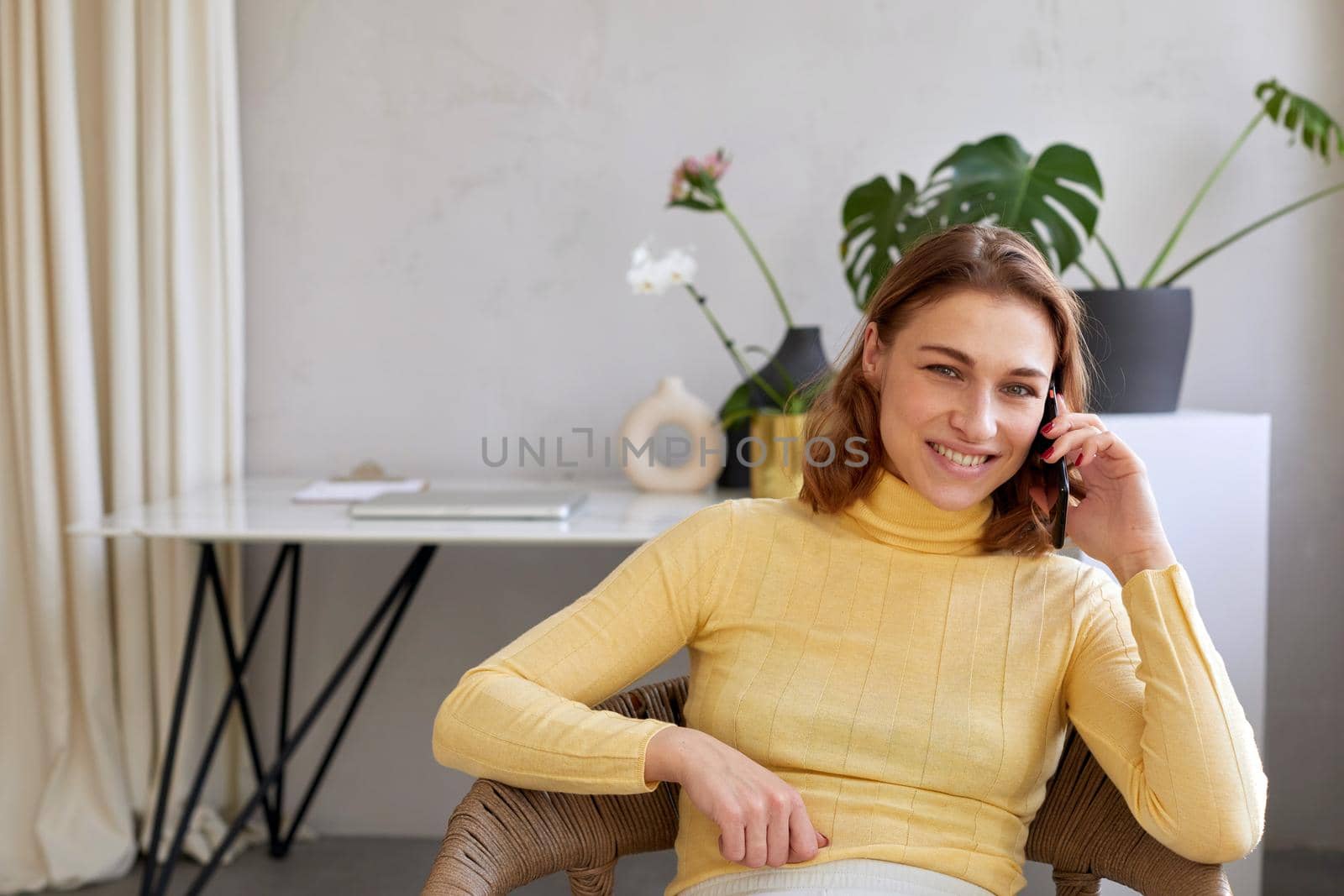 Happy woman in yellow turtleneck winking gesture while sitting on chair and talking on mobile phone in light workspace