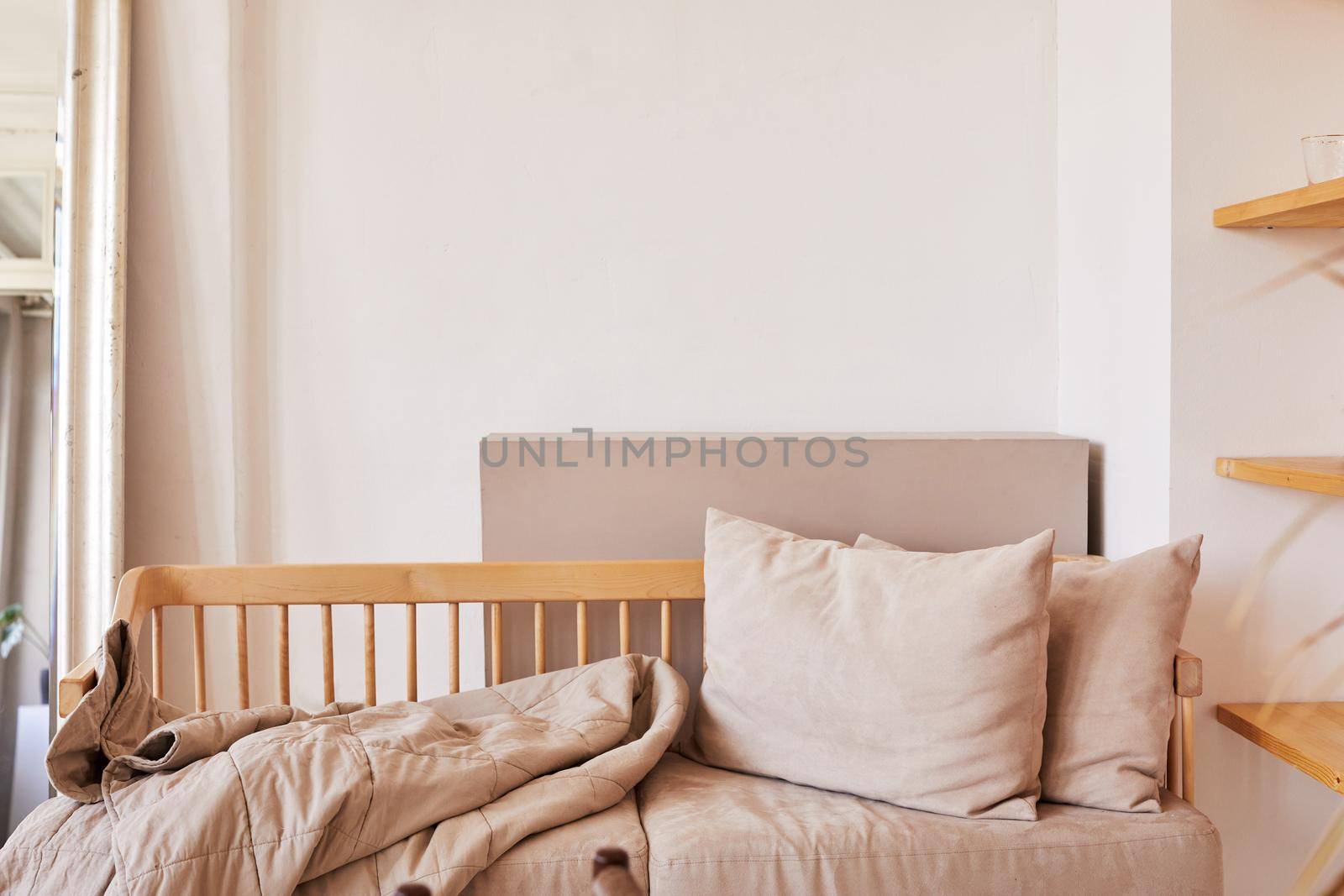 Fragment of home interior in natural pastel tones with wooden couch with soft beige cushions and coverlet placed against white wall