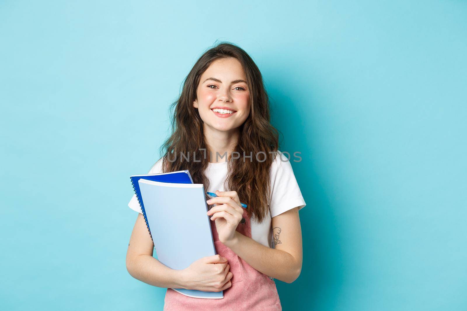 Happy young female student holding notebooks from courses and smiling at camera, standing in spring clothes against blue background by Benzoix