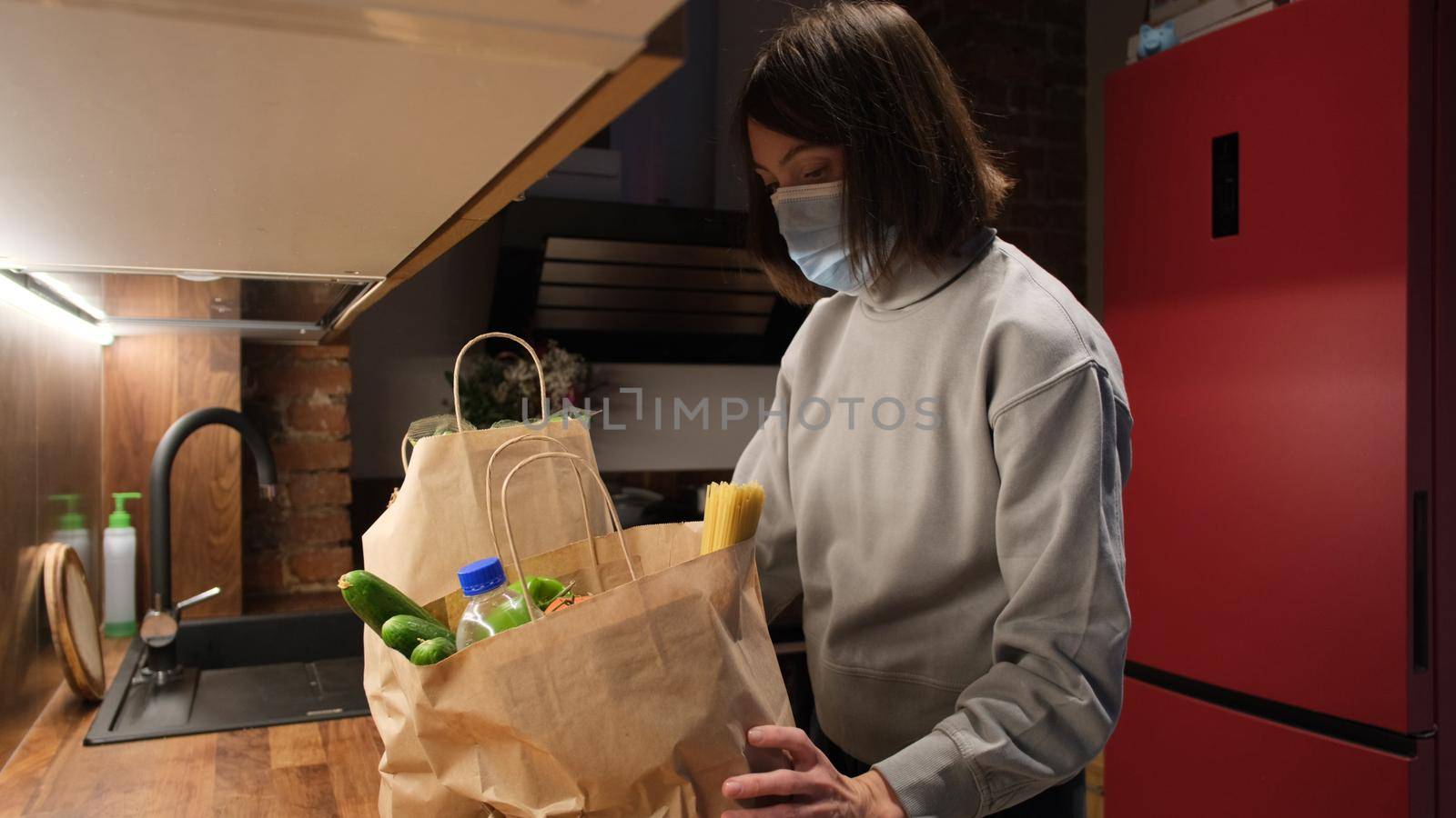 Young short haired housewife puts large paper bags with products on countertop in protective mask in kitchen at home