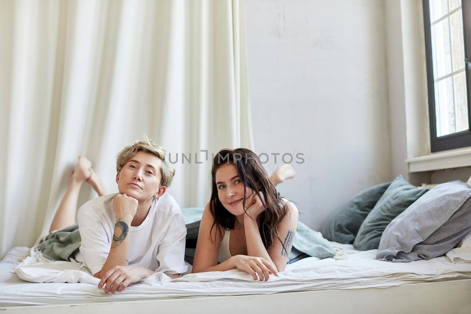 Young women leaning on hands and looking at camera while chilling on bed in morning in cozy bedroom at home