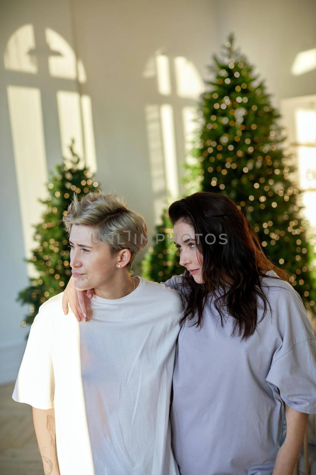 Same sex partners embracing each other while standing against coniferous tree decorated with garlands and looking away with interest
