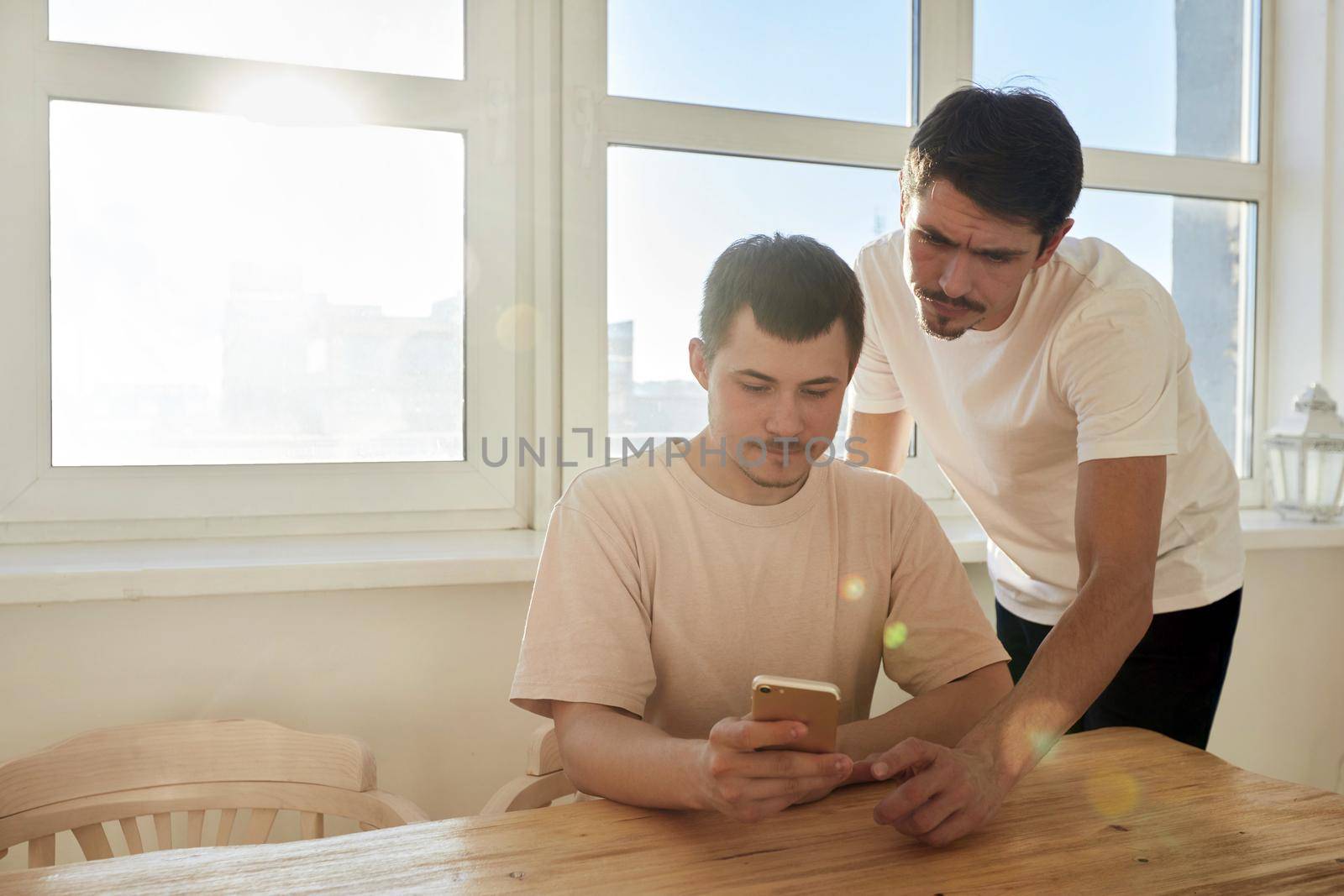 Boyfriends in similar clothes resting near table and browsing social media on smartphone against window with bright sunlight at home
