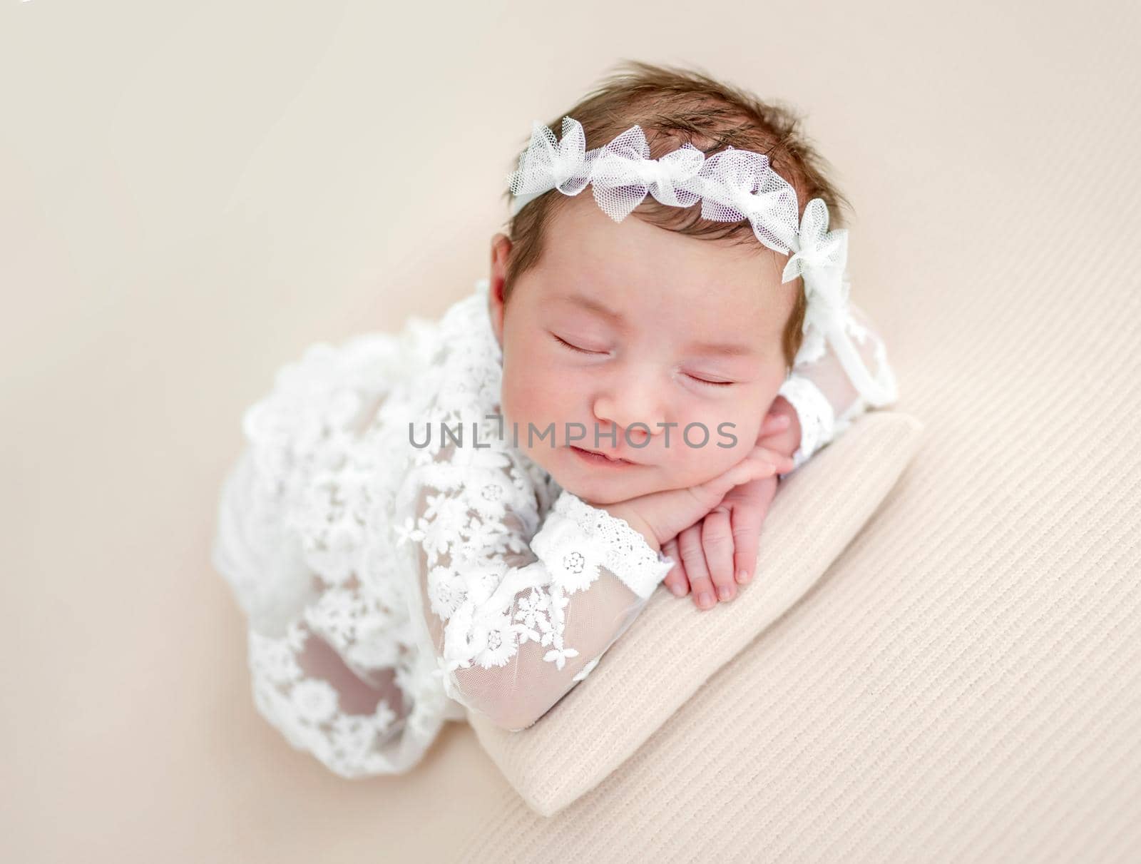 Closeup portrait of adorable newborn baby girl wearing beautiful white dress and wreath lying on her tummy and sleeping in studio. Cute infant child napping holding hands under cheeks