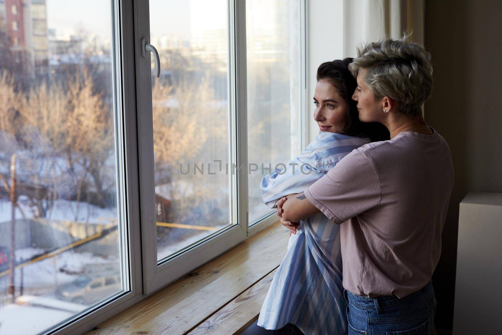 Back view of woman with short hair embracing girlfriends and looking out window while resting at home on winter day