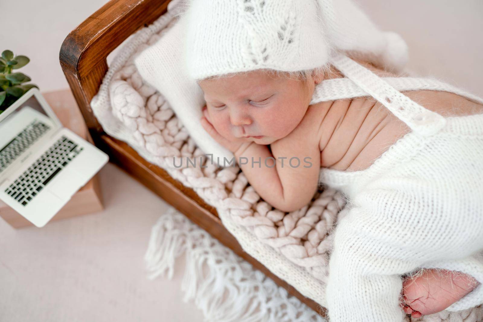 Newborn baby boy sleeping in tiny bed wearing knitted white costume and hat. Infant kid studio portrait with tiny toy laptop