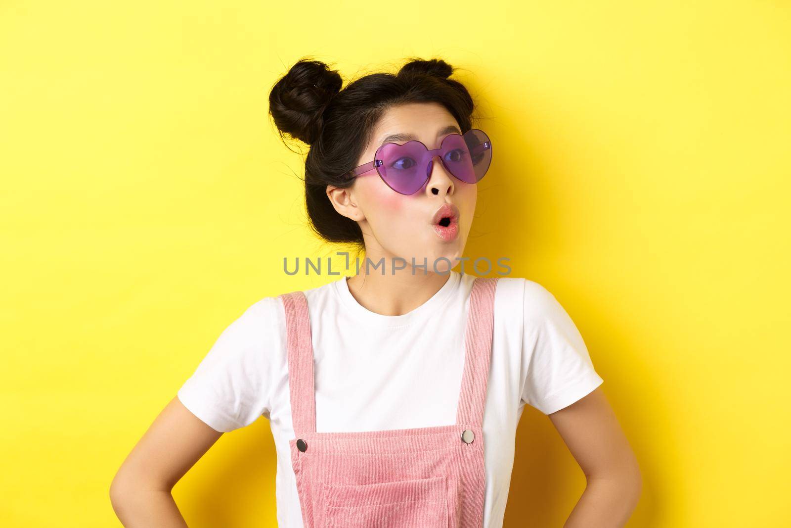 Summer and fashion concept. Close-up of surprised asian teen girl in heart-shaped sunglasses looking right side with excited face, standing on yellow background by Benzoix