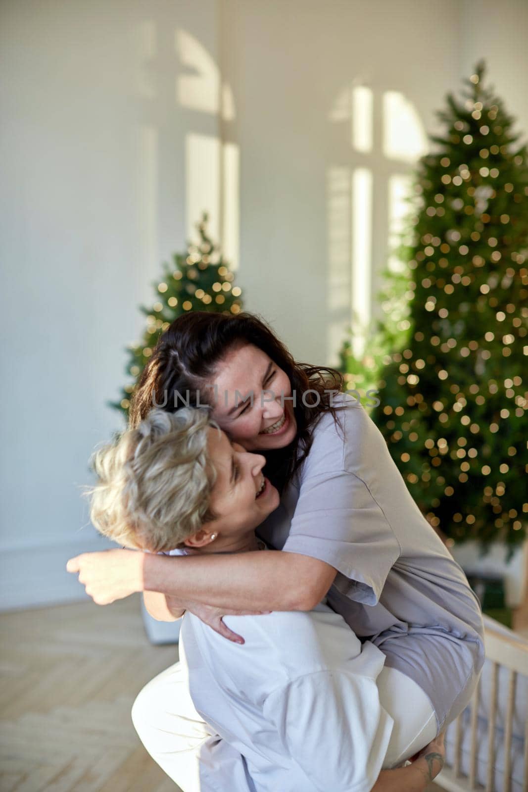 Cheerful young women laughing and embracing each other while celebrating Christmas in cozy room at home