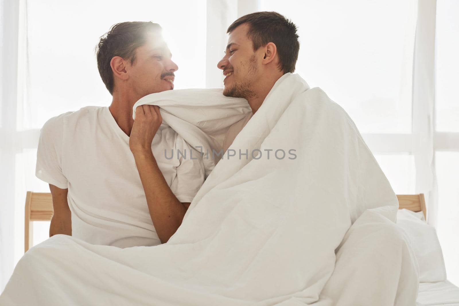 Happy young men wrapping in warm blanket and smileing at each other while resting on comfortable bed in morning in cozy bedroom
