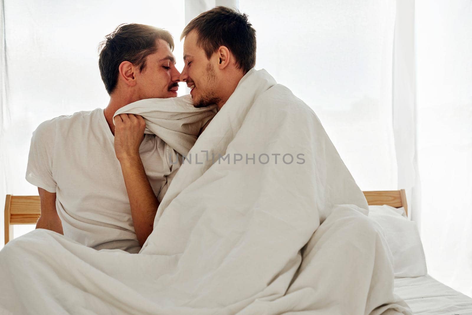 Happy young men wrapping in warm blanket and touching noses while resting on comfortable bed in morning in cozy bedroom