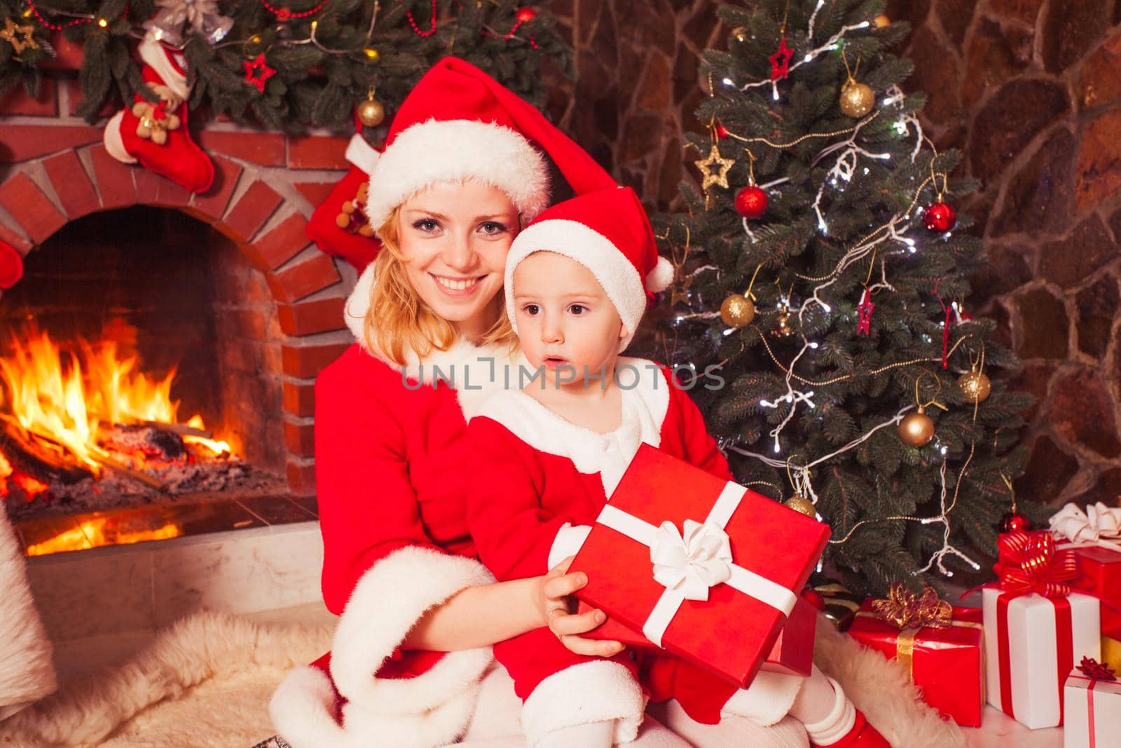 Mother and son are sitting near fireplace and christmas tree. Family look into a gift box.