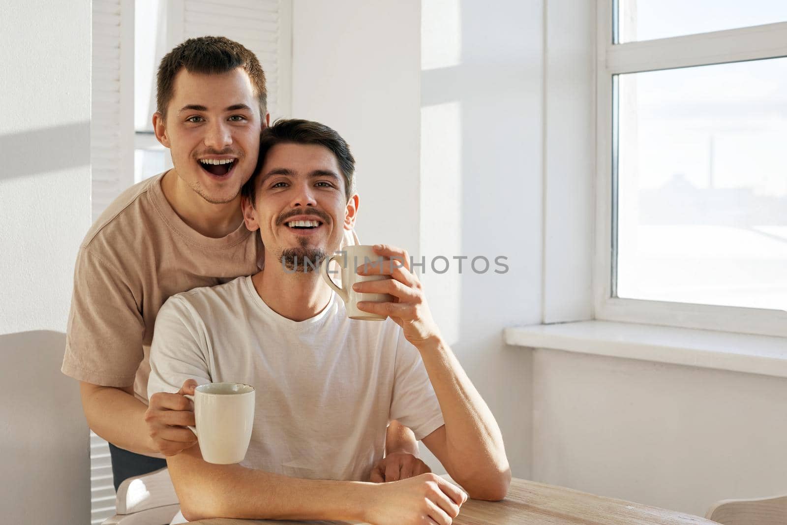 Cheerful young men with mugs of hot drinks smiling and looking at camera while sitting at table in morning at home