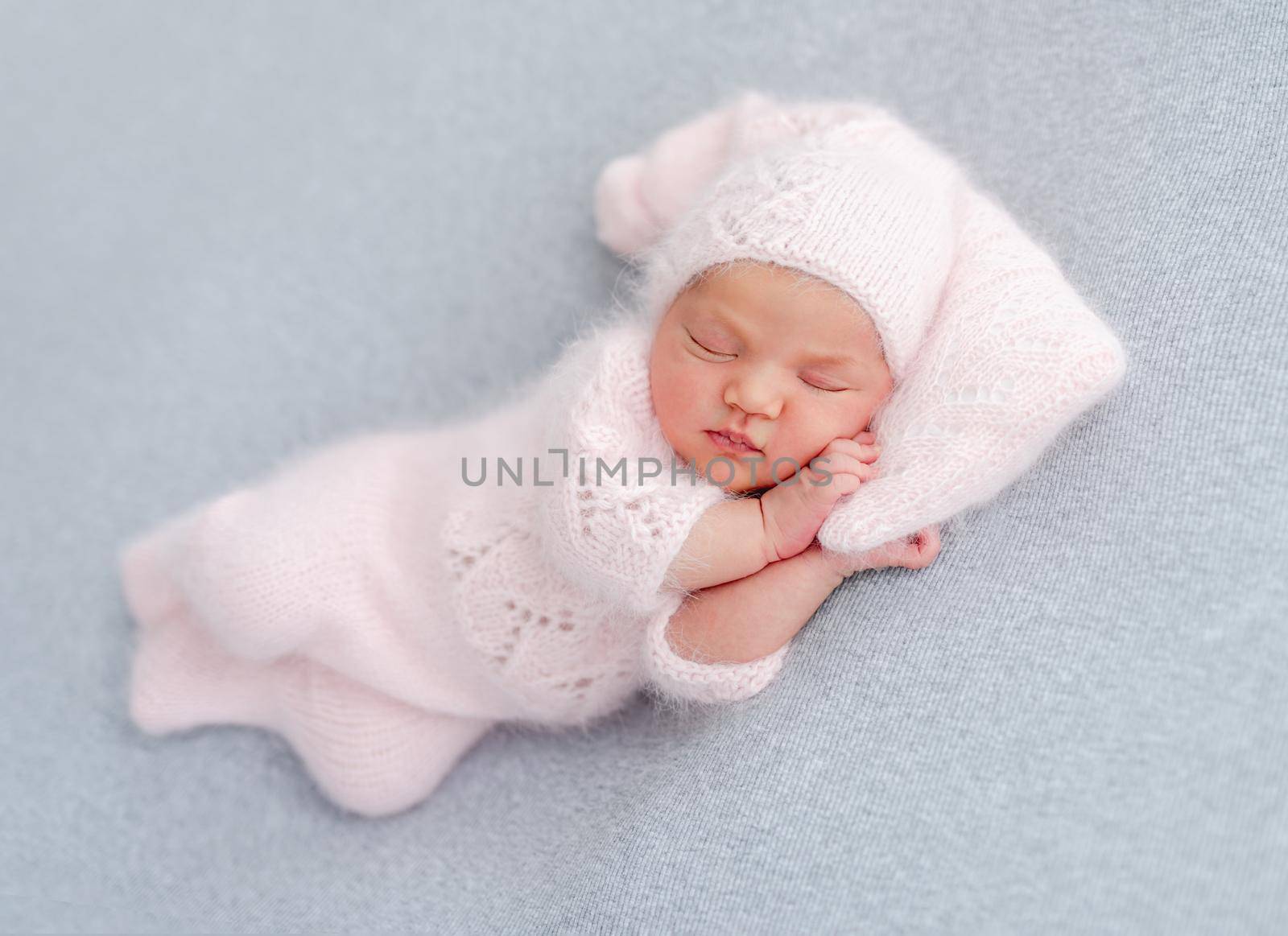 Cute newborn baby girl in knitted hat and costume sleeping on grey background and holding her hands under cheek. Adorable infant child napping during studio photoshoot