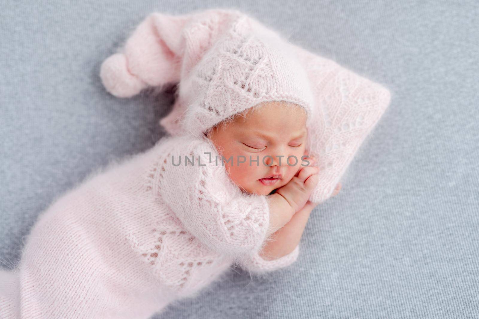 Cute newborn baby girl in knitted hat and costume sleeping on grey background and holding her hands under cheek. Adorable infant child napping during studio photoshoot. Little kid closeup portrait