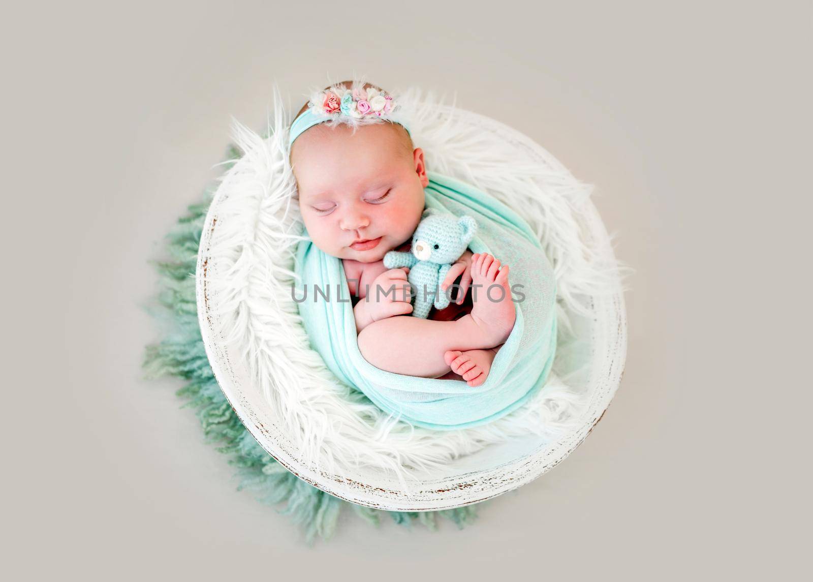 Adorable newborn baby girl lying on her back in the cocoon in white basin, sleeping and holding knitted teddy bear in her hands. Sweet infant child napping with toy during studio photoshoot