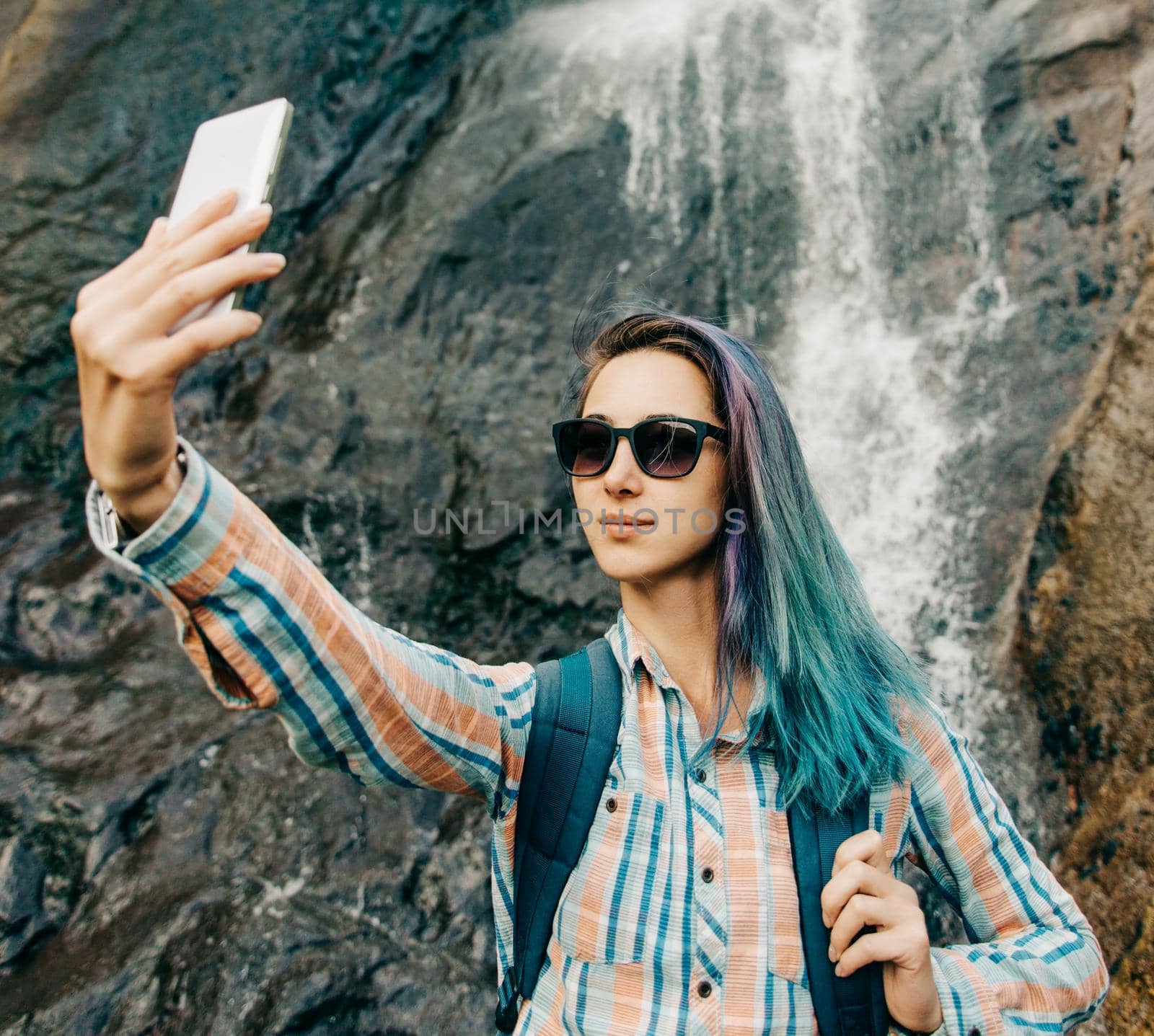 Traveler backpacker young woman taking selfie with smartphone on background of waterfall in summer outdoor.