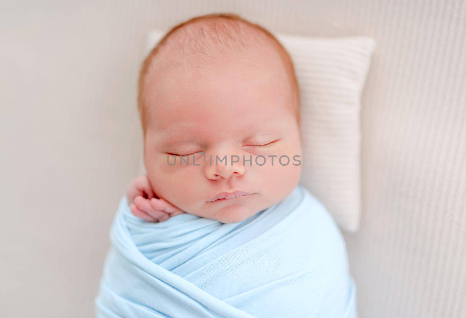 Portrait of newborn baby boy swaddled in light blue fabric sleeping on white background and holding tiny hands under his cheeks. Adorable infant child napping during studio photoshoot