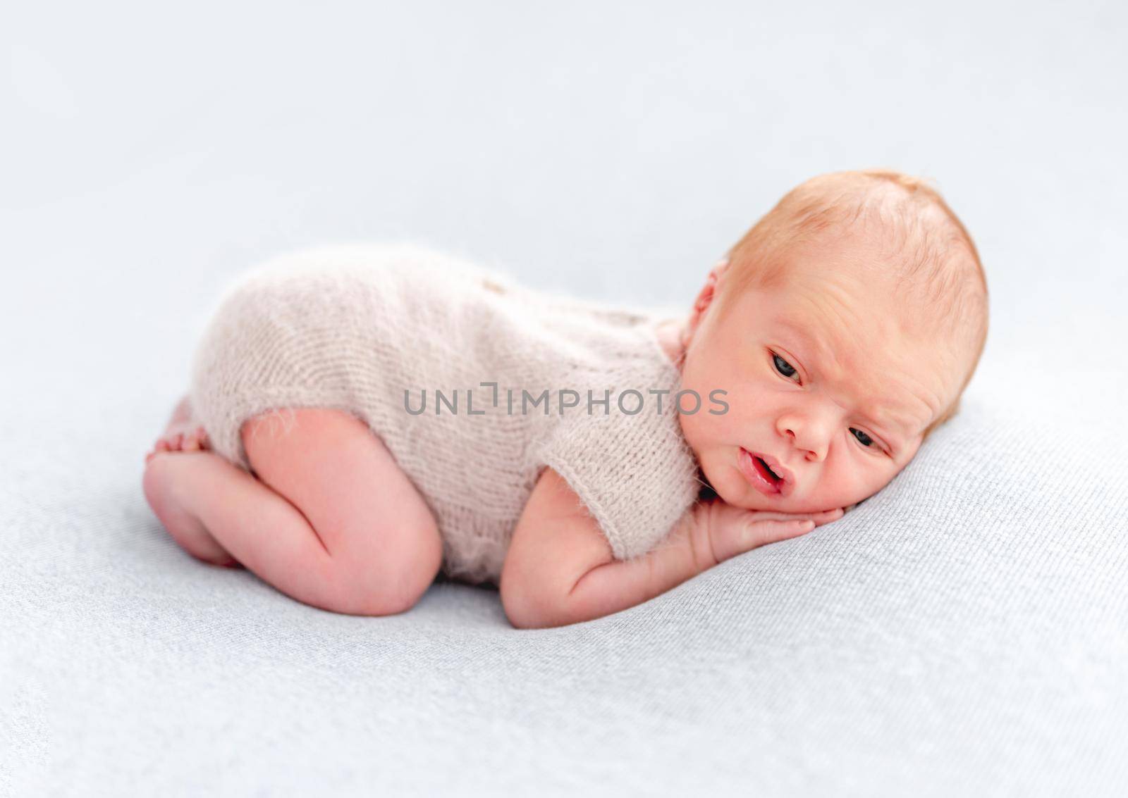 Newborn baby boy wearing knitted beige costume lying on his tummy and holding tiny hands under his cheeks. Adorable infant child resting and looking at the camera