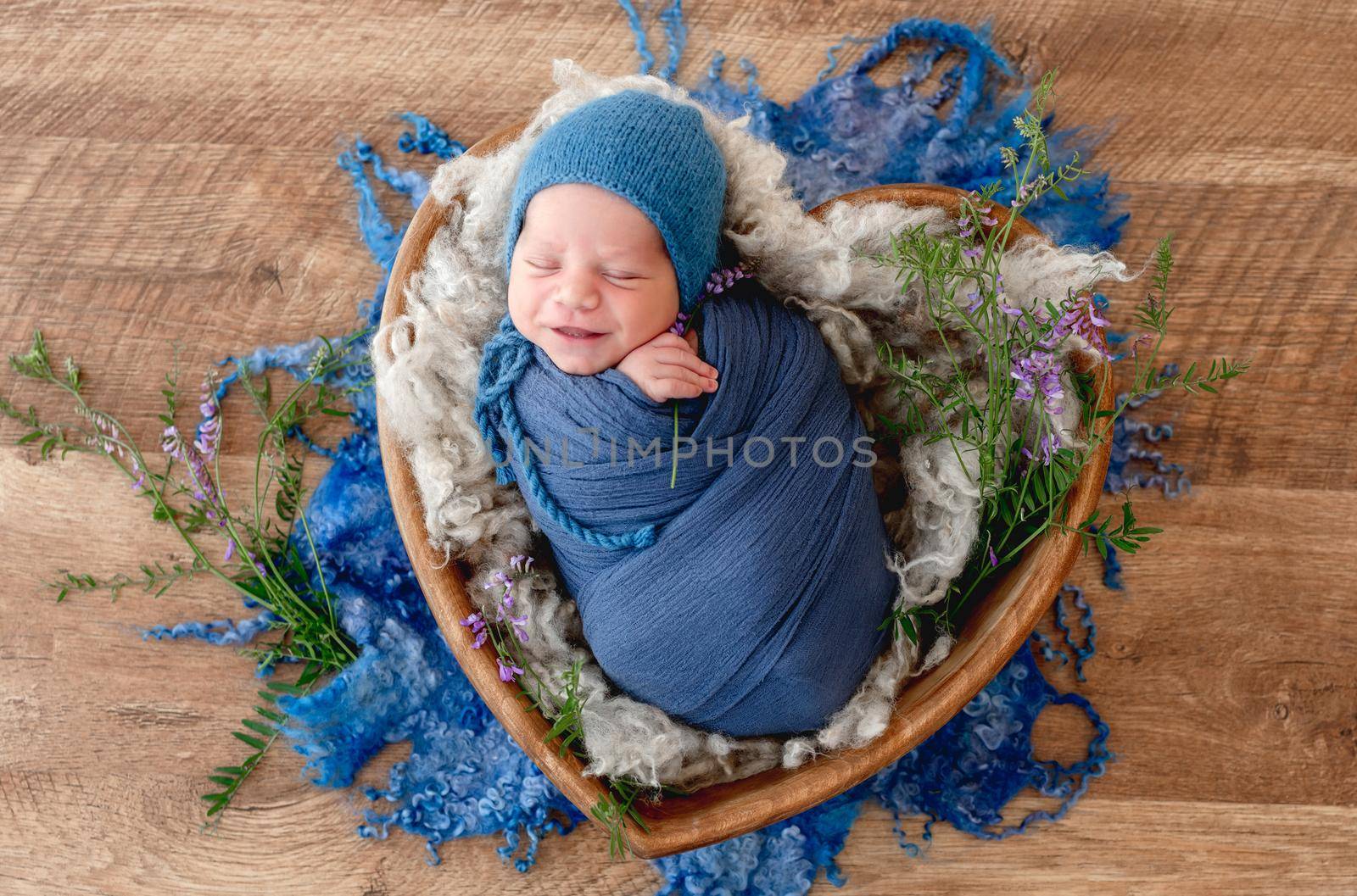 Adorable newborn baby boy swaddled in blue fabric holding lavander flower in his hands and smiling during sleeping. Cute infant kid napping in fur in wooden heart bed during studio photoshoot.