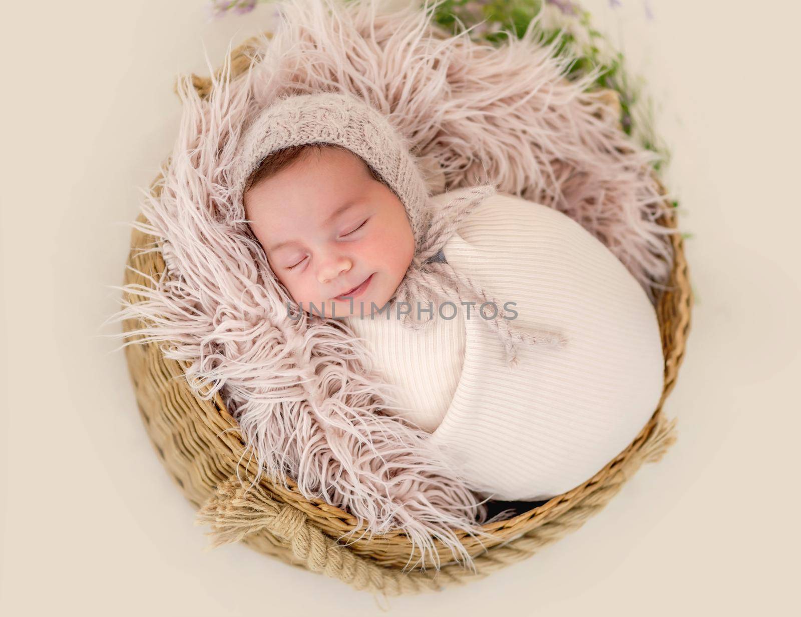 Portrait of beautiful newborn baby girl swaadled in fabric and wearing knitted hat sleeping in basket with fur during studio photoshoot. Cute infant child napping