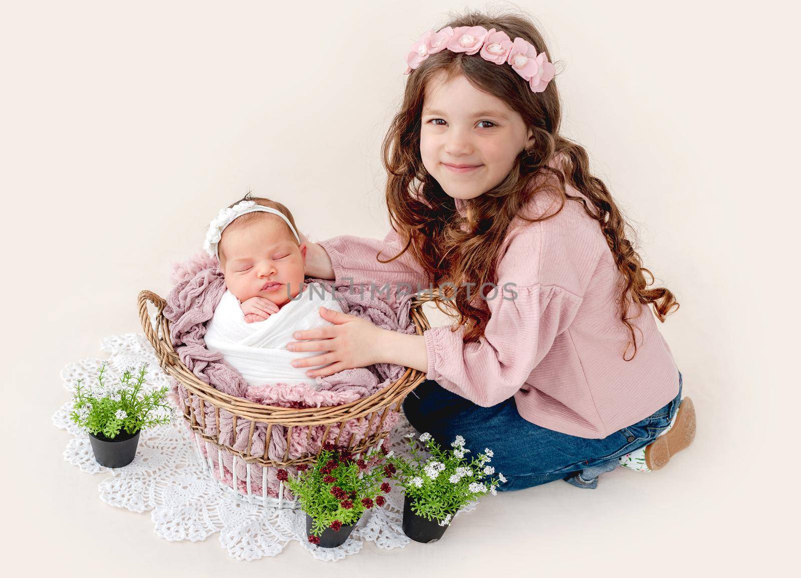Beautiful little girl smiling and sitting with her swaddled beautiful newborn baby sister sleeping in the basket. Adorable napping kid with her sibling