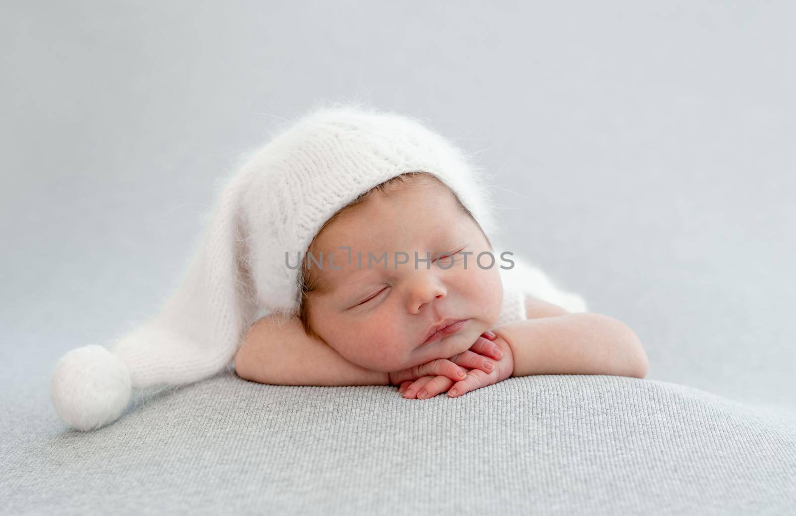Newborn baby boy sleeping wearing knitted white hat and holding his hands under cheeks. Infant kid studio portrait