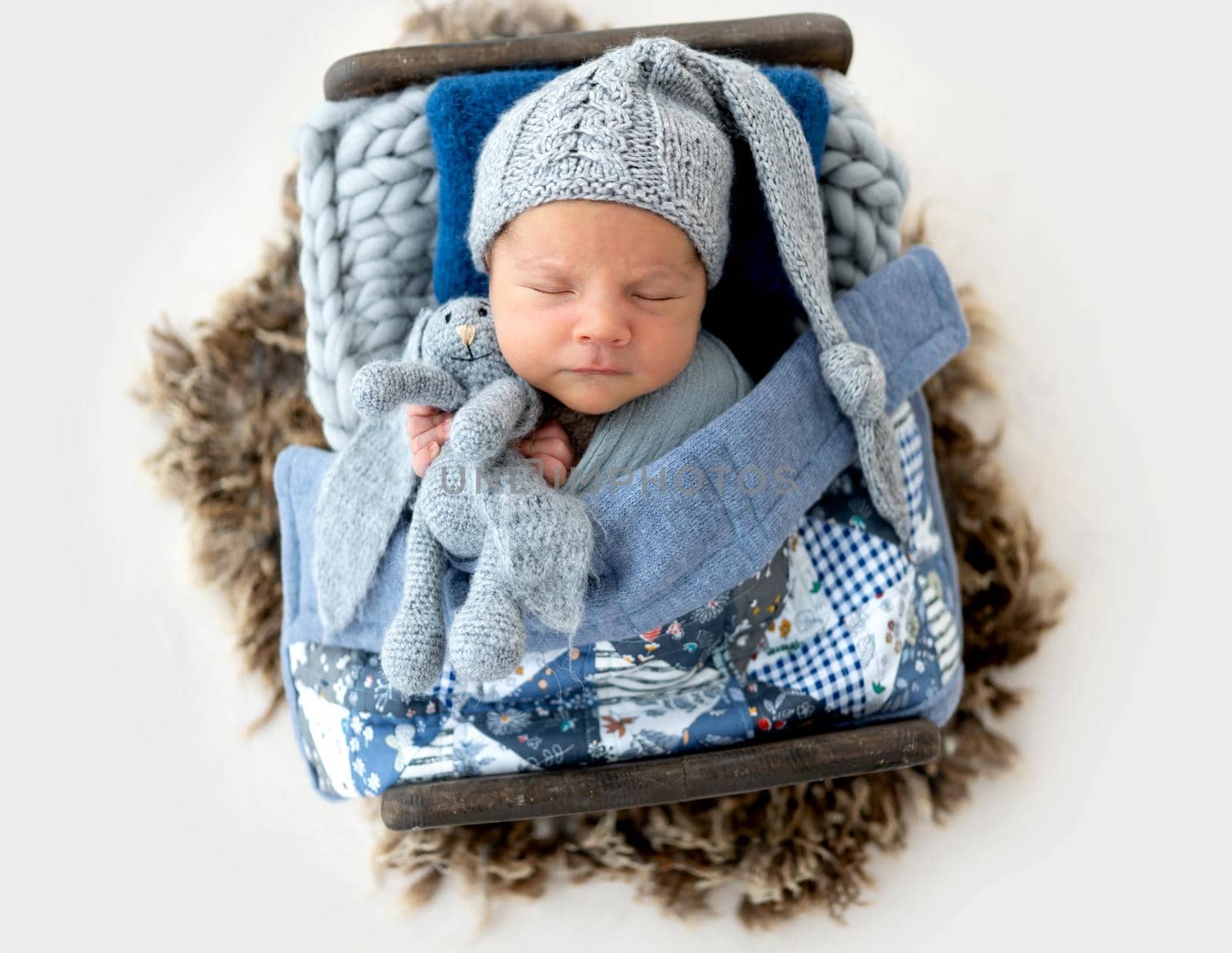 Adorable newborn baby boy swaddled in fabric and wearing cute knitted hat sleeping in wooden stylized bed and holding bunny toy in tiny hands. Infant child napping indoors