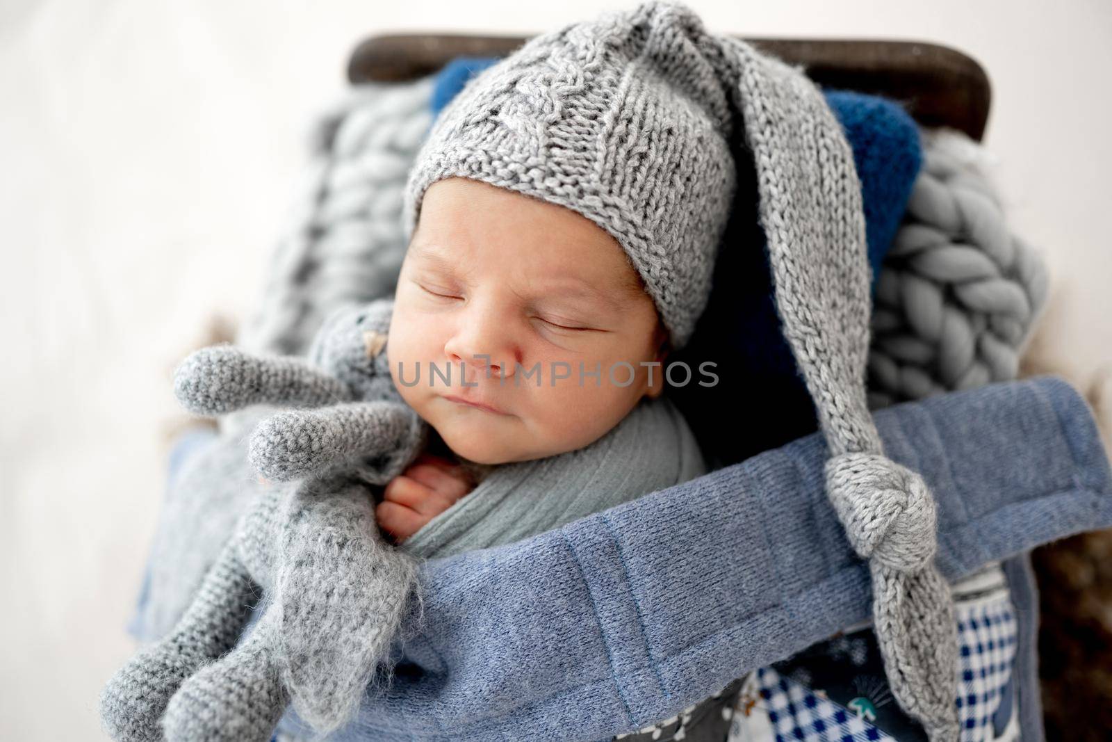 Adorable newborn baby boy wearing cute knitted hat sleeping and holding bunny toy in tiny hands during studio photoshoot. Infant child napping indoors