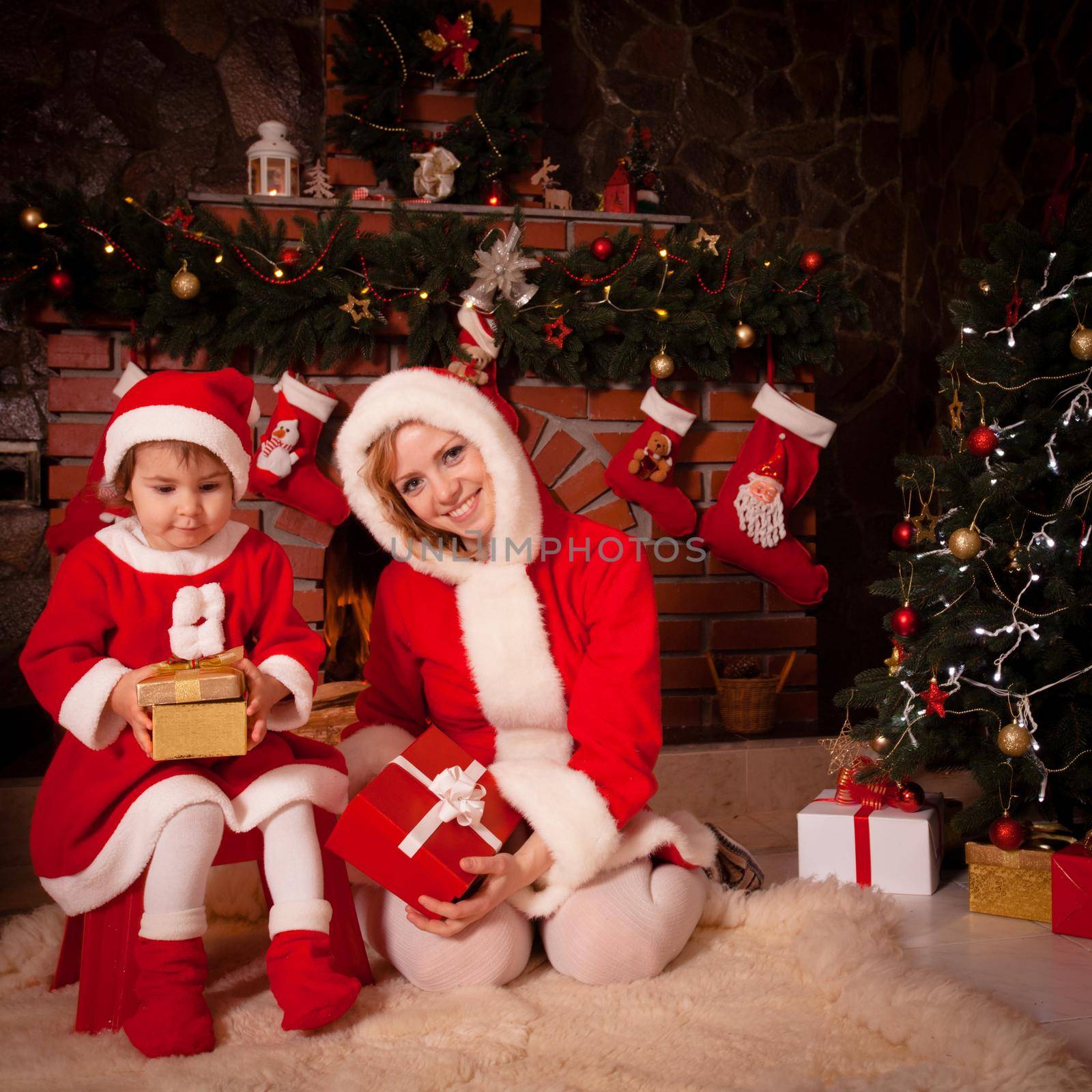 Mother and daughter are sitting near fireplace and christmas tree with gift boxes.