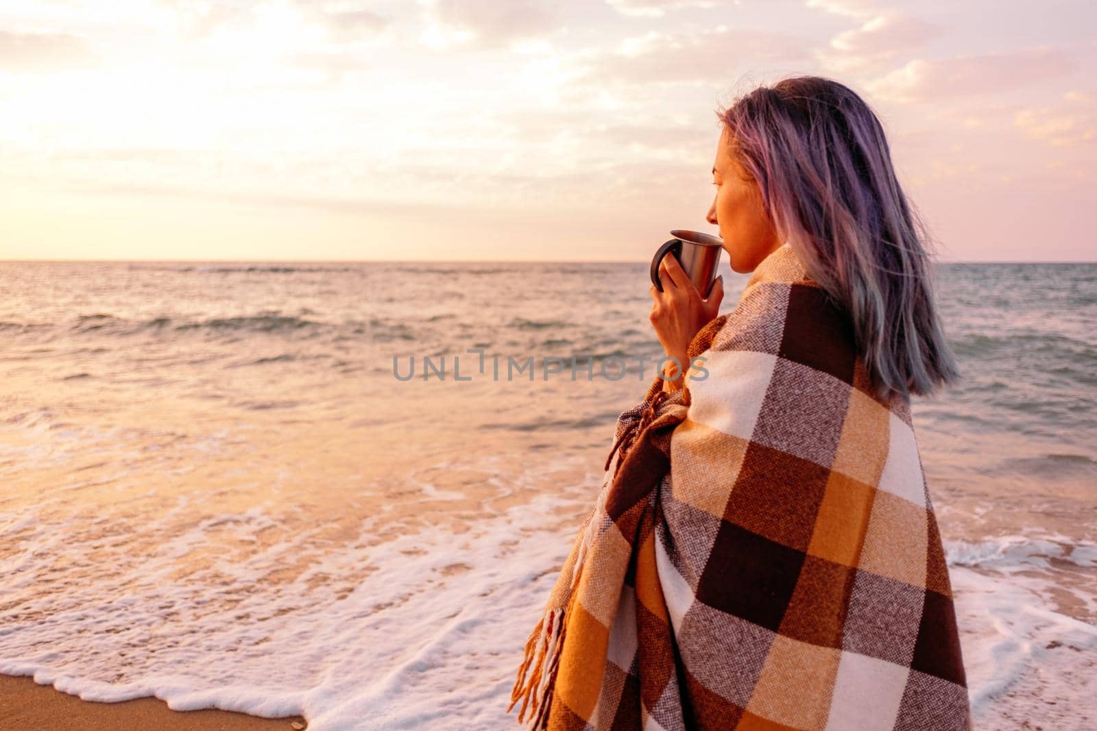 Young woman with cup of beverage resting on shore near the sea at sunset. Summer beach vacations.