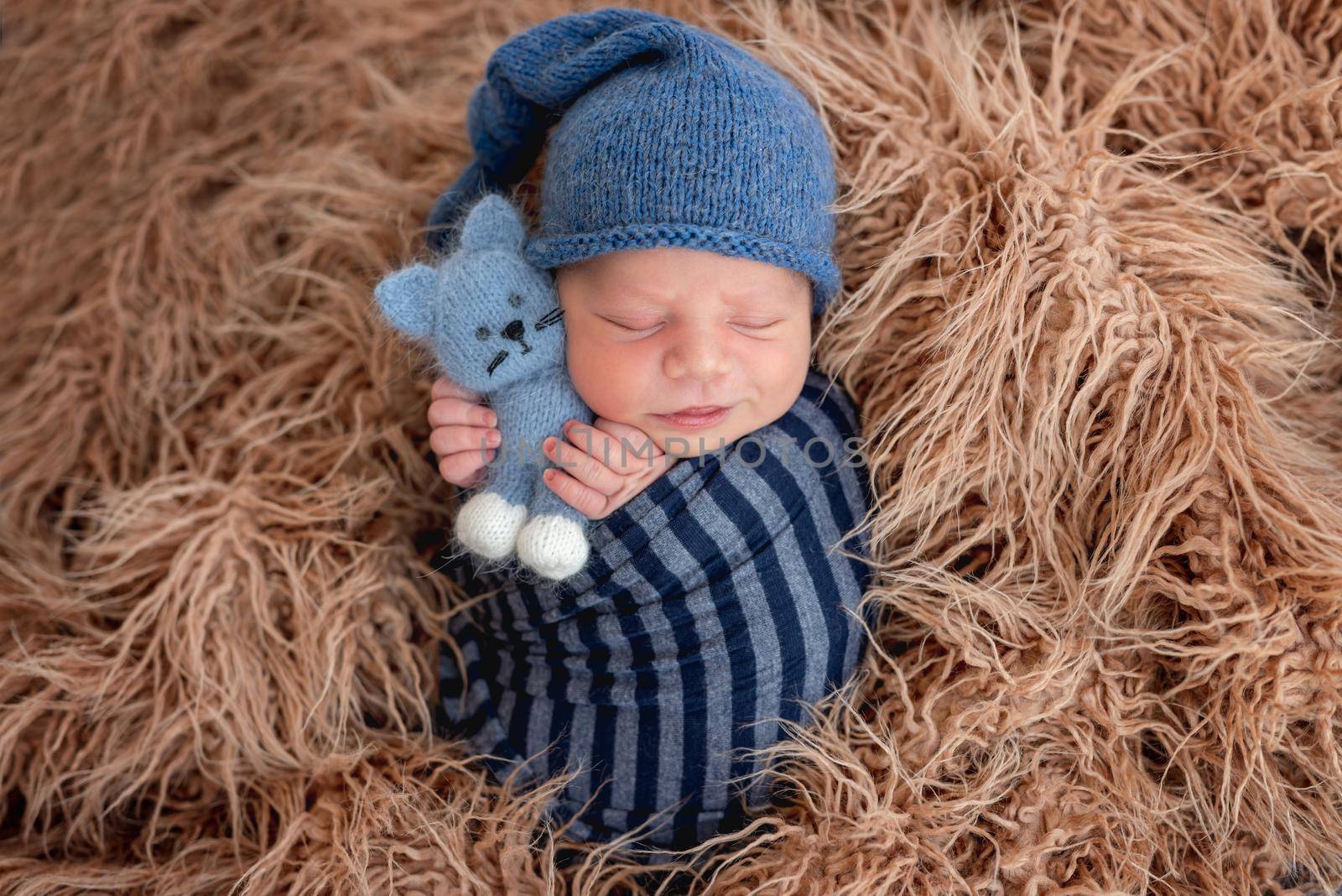 Adorable newborn baby boy swaddled in toby fabric hugging knitted toy and smiling during sleeping. Cute infant kid wearing hat napping in fur during studio photoshoot.