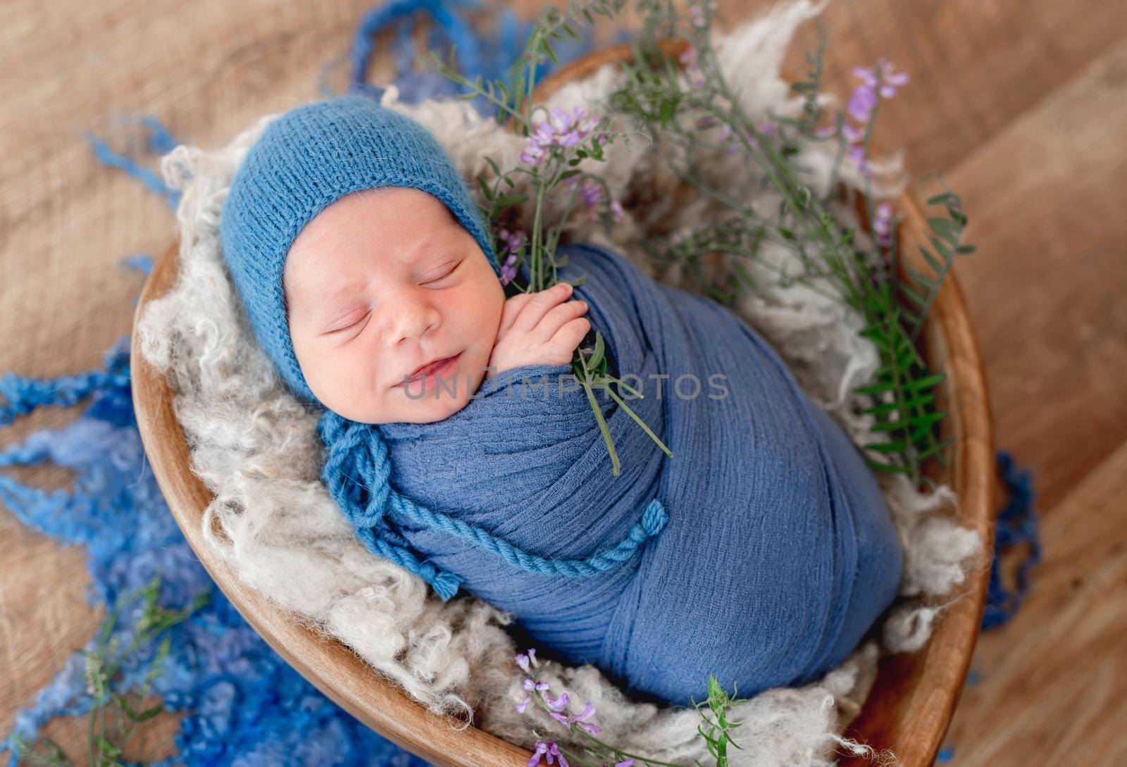 Adorable newborn baby boy swaddled in blue fabric holding purple flower in his hands smiling during sleeping. Cute infant kid wearing knitted hat in fur in wooden heart bed during studio photoshoot