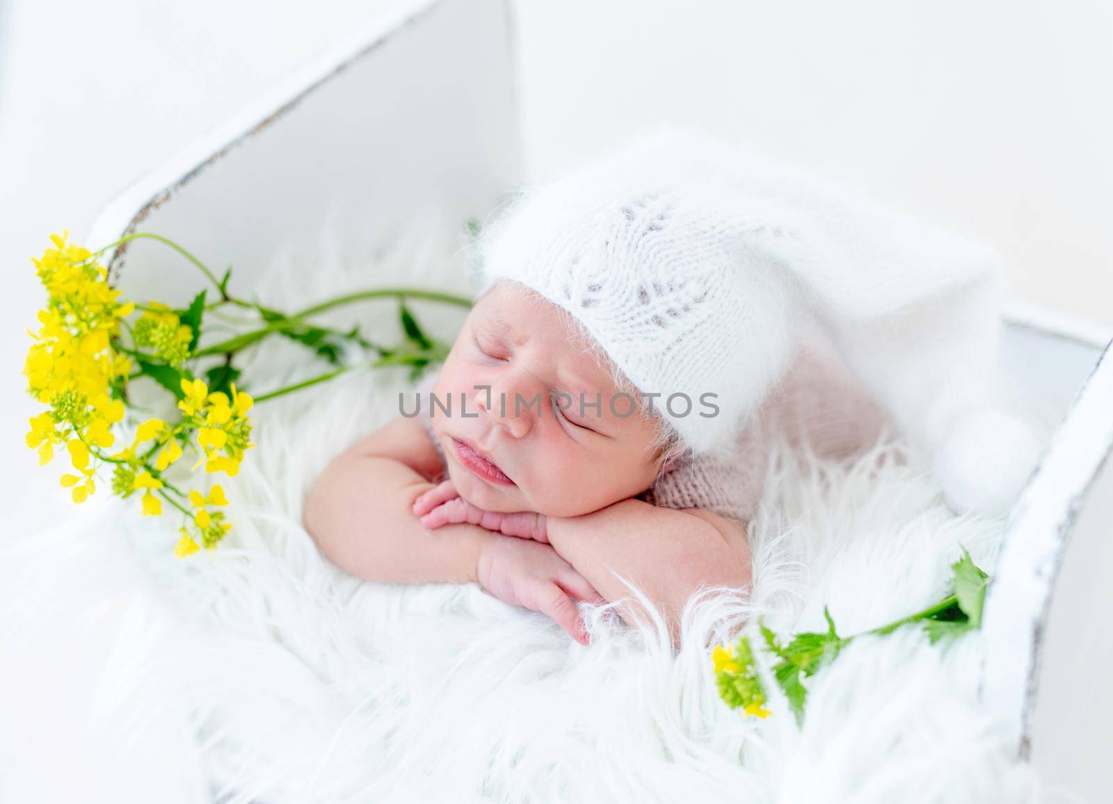 Portrait of adorable newborn baby boy sleeping on his tummy and holding his hands under cheeks in white wooden bed with yellow flowers. Cute infant kid wearing knitted hat napping