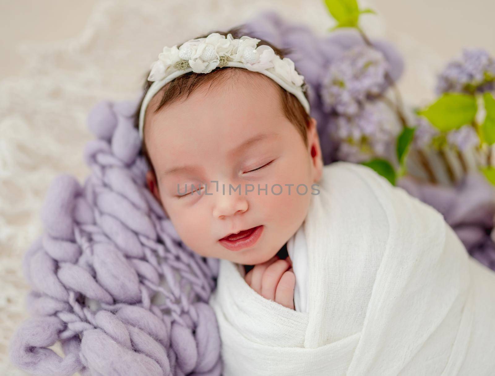 Portrait of little beautiful newborn baby girl swaadled in fabric and wearing wreath with flowers sleeping and smiling during studio photoshoot. Cute infant child kid napping on knitted blanket