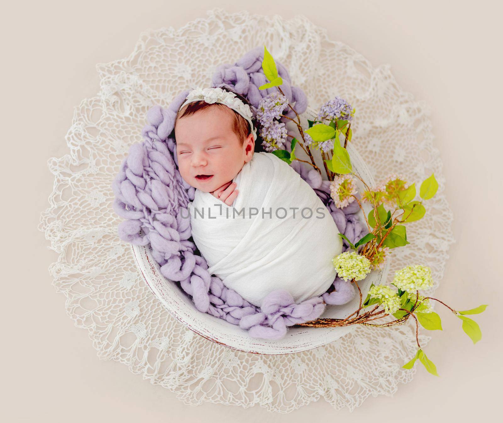 Portrait of little beautiful newborn baby girl swaadled in fabric and wearing wreath with flowers sleeping and smiling in basin with decoration during studio photoshoot. Cute infant child