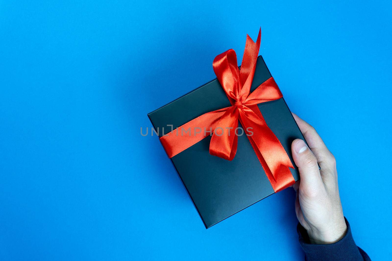 Right mans hand gifts a present black box with red ribbon on classic blue background, the color of the year 2020. Enough place for text.